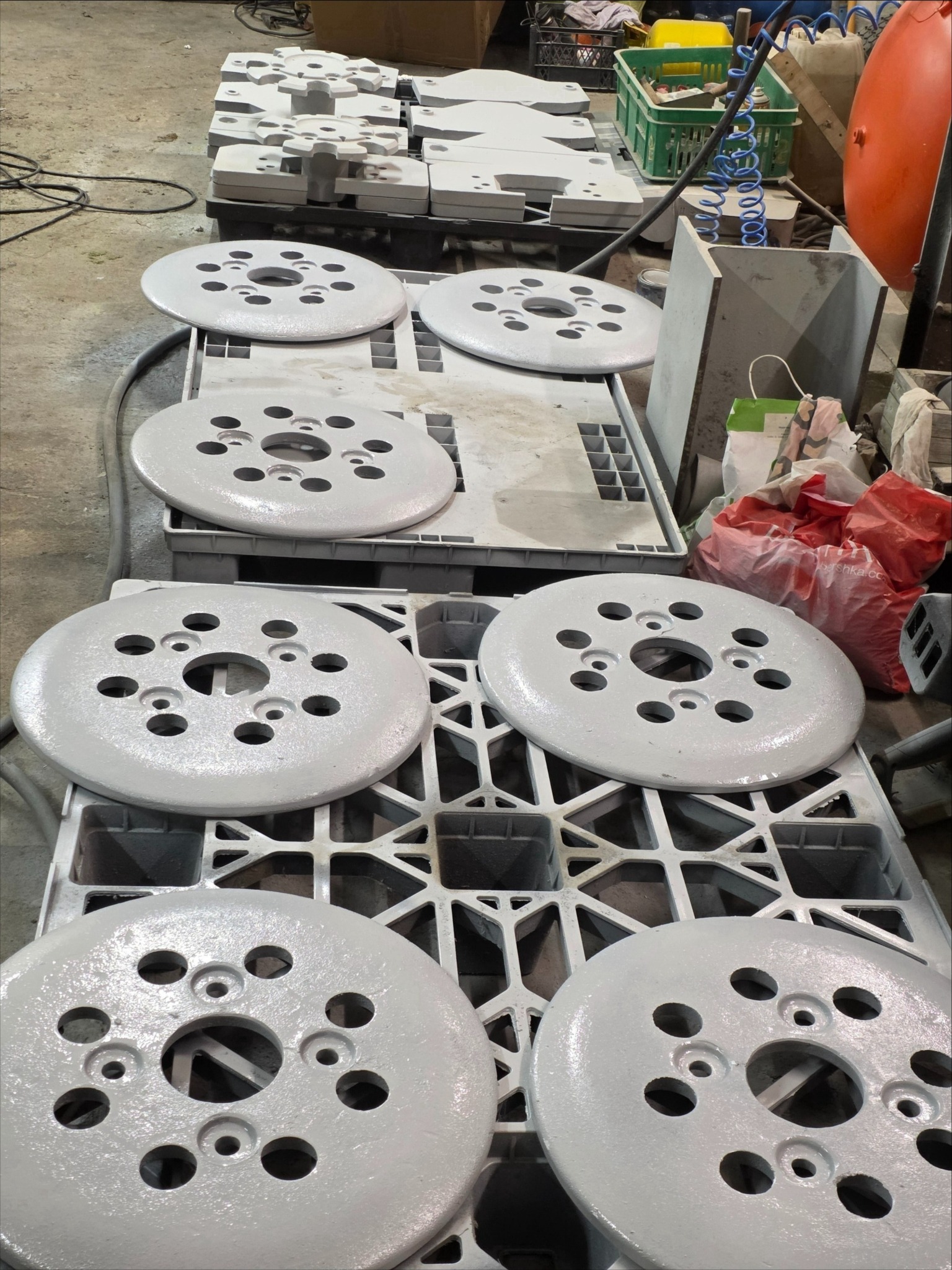 Metalowe elementy po obróbce, pomalowane na szaro, ułożone na paletach. Widoczne ślady po procesie czyszczenia. Elementy okrągłe z otworami.