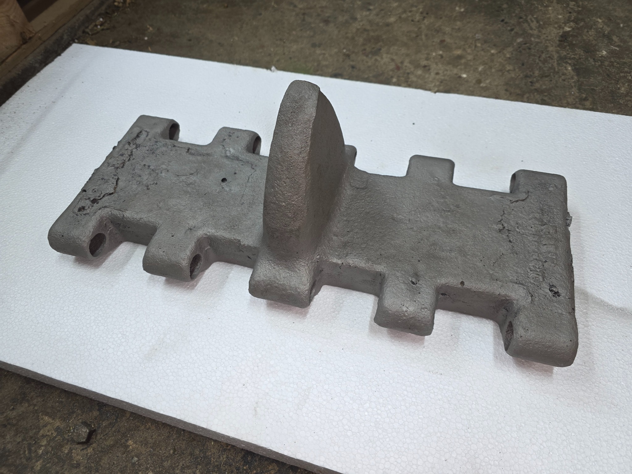 Oczyszczony ogniwo gąsienicy po obróbce, leżący na białym podłożu. Widoczne ślady wcześniejszej korozji i nierówności powierzchni. Szary odcień metalu.