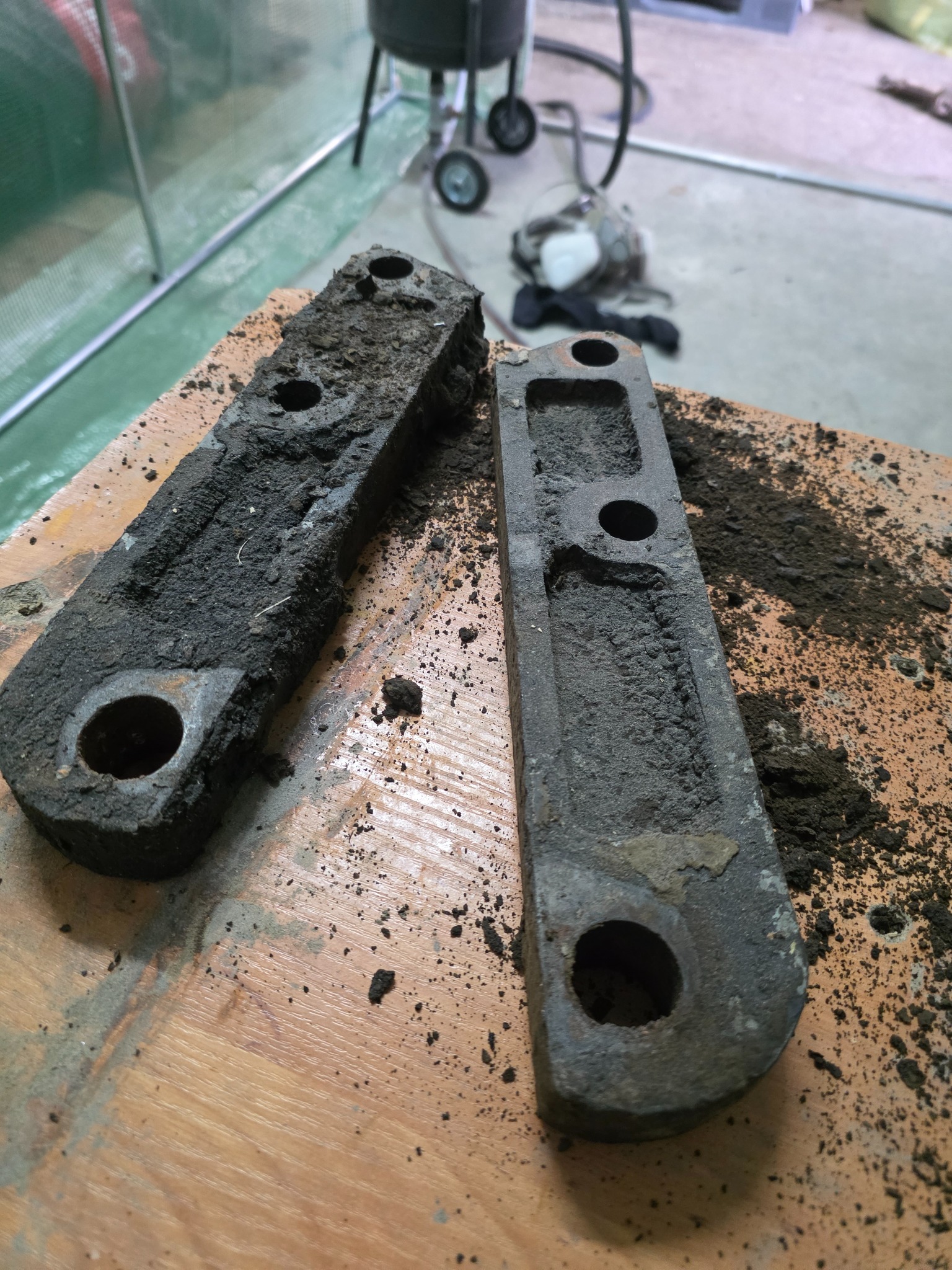 Dwie metalowe części, jedna pokryta grubą warstwą brudu, druga oczyszczona, leżą na drewnianym stole. W tle widoczny sprzęt do czyszczenia.