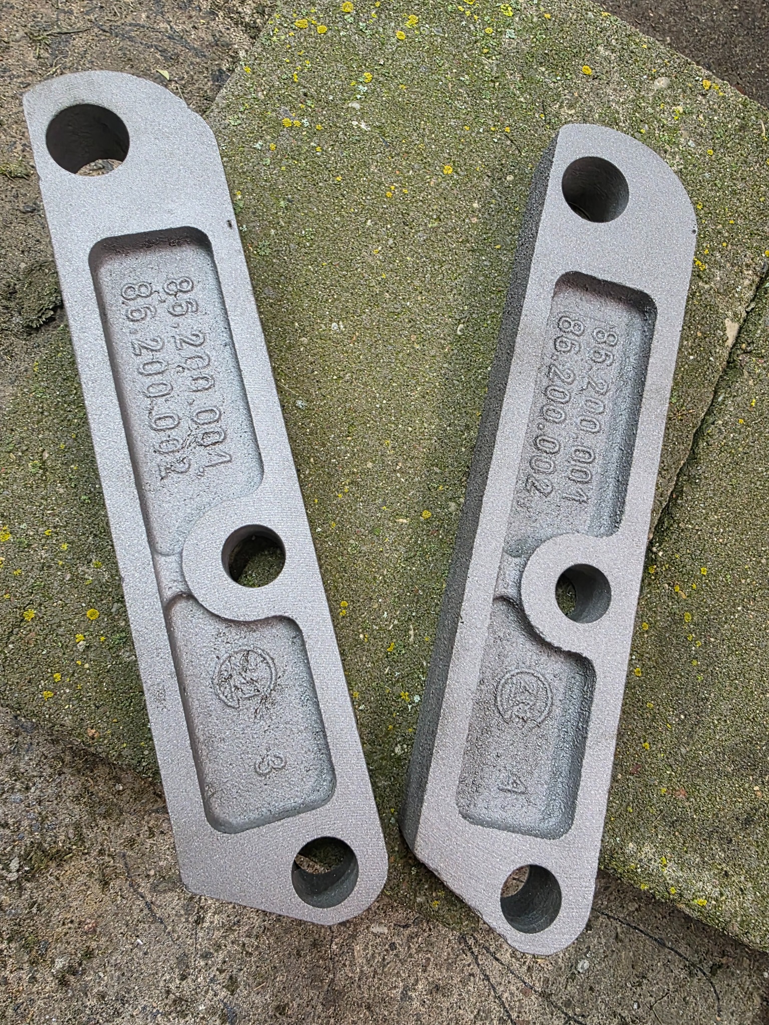Dwa metalowe elementy po obróbce, leżące na szorstkiej powierzchni. Widoczne numery seryjne i otwory montażowe. Szary odcień metalu, chropowata faktura.