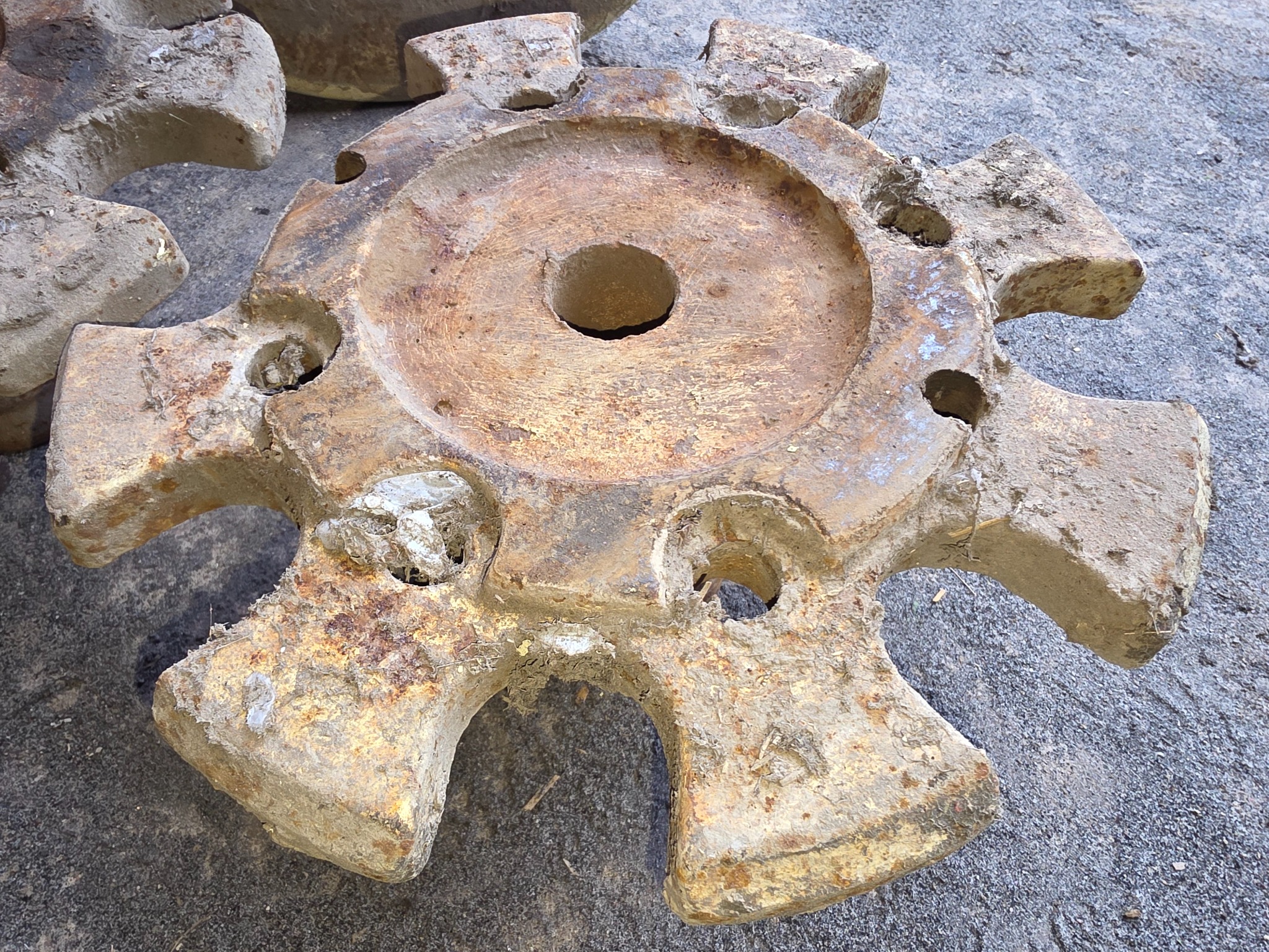 Zardzewiały, okrągły element maszyny z otworami i wypustkami, pokryty resztkami po obróbce, leżący na szarym podłożu. Widoczne ślady korozji i zabrudzenia.