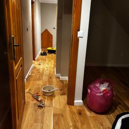 Paweł Kwaśny - Korytarz z nową, błyszczącą podłogą z paneli drewnianych. Widoczne narzędzia, wiadro i worek na śmieci sugerują prace wykończeniowe w toku. Ciepłe oświetlenie podkreśla naturalny rysunek drewna.