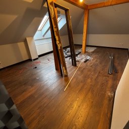 Paweł Kwaśny - Podłoga z paneli w odcieniach brązu w pokoju na poddaszu, widoczne listwy przypodłogowe, narzędzia i ramy drzwiowe. Ukończony etap prac wykończeniowych.