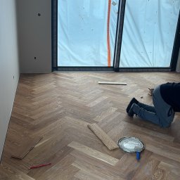 Paweł Kwaśny - Układanie parkietu w jodełkę: widok z góry na częściowo ułożoną podłogę, narzędzia pracy i osobę w trakcie montażu. Okno zasłonięte folią ochronną.