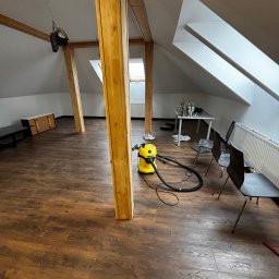 Paweł Kwaśny - Poddasze z drewnianymi belkami i nową, ciemną podłogą. Żółty odkurzacz stoi na panelach. Widoczne okna dachowe i białe ściany. Ustawione krzesła i stół.