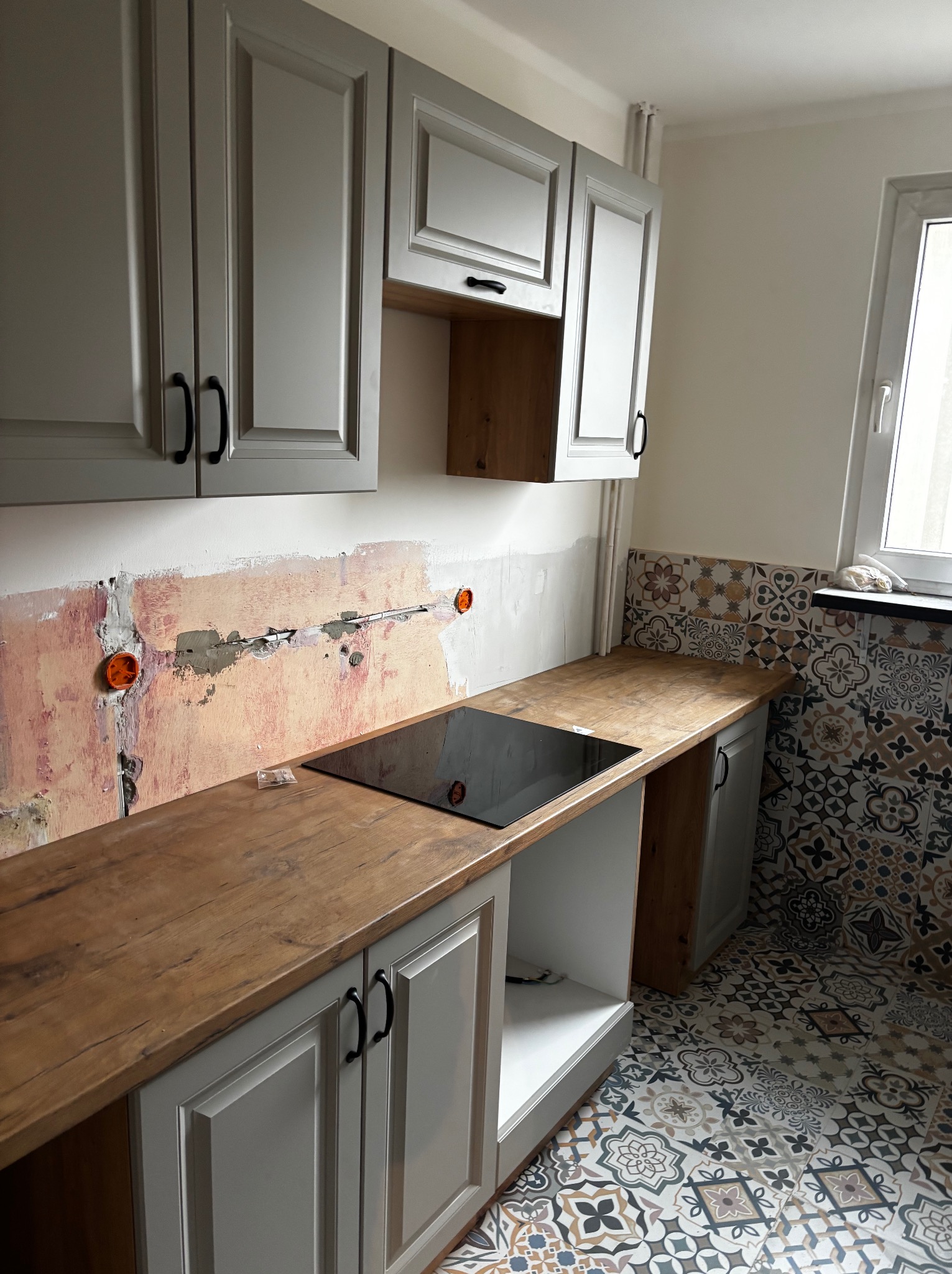 Kuchnia w trakcie remontu: szare szafki z drewnianym blatem, płyta indukcyjna, fragment ściany z instalacją elektryczną i wzorzyste płytki podłogowe.