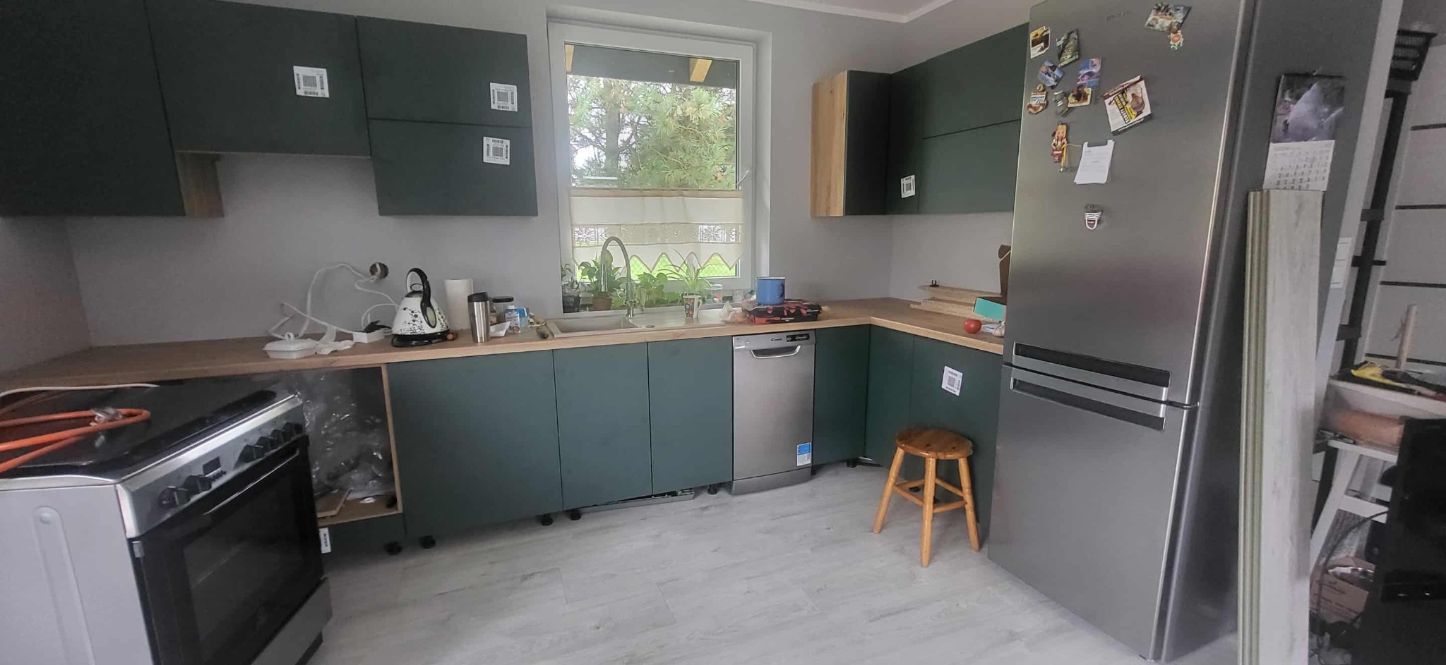 Nowoczesna kuchnia z zielonymi szafkami i drewnianym blatem. Widoczny sprzęt AGD: lodówka, zmywarka i kuchenka. W tle okno z widokiem na zieleń.