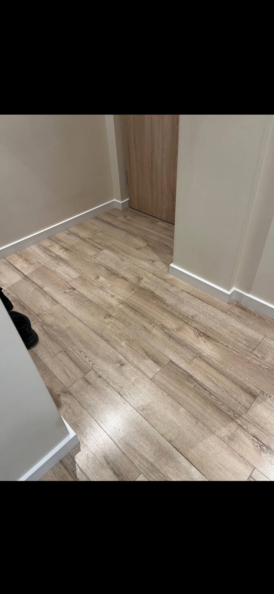 Jasne panele podłogowe ułożone wzdłuż ściany z białą listwą przypodłogową i drewnianymi drzwiami w tle. Widok z góry, neutralne oświetlenie.