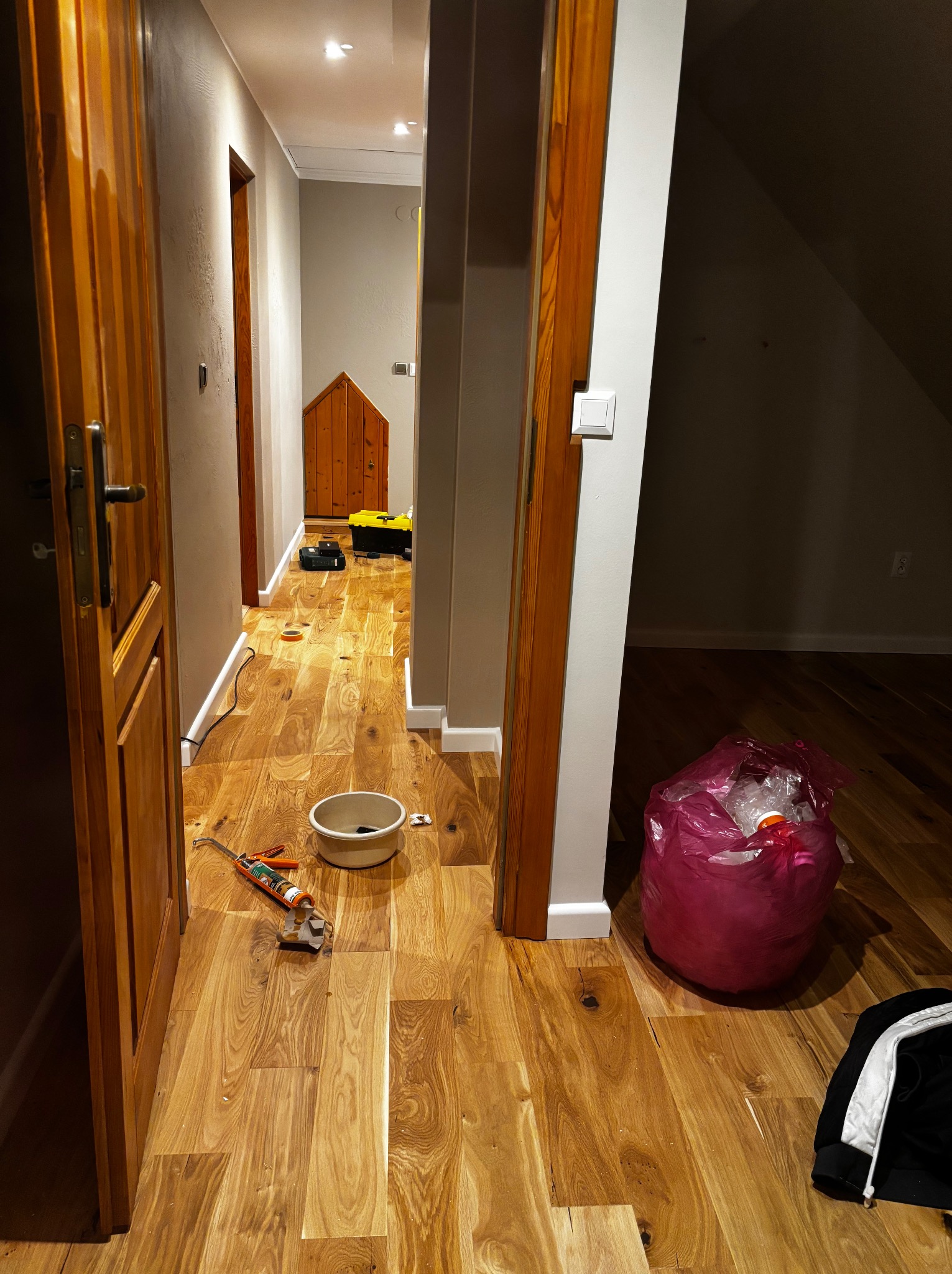 Korytarz z nową, błyszczącą podłogą z paneli drewnianych. Widoczne narzędzia, wiadro i worek na śmieci sugerują prace wykończeniowe w toku. Ciepłe oświetlenie podkreśla naturalny rysunek drewna.