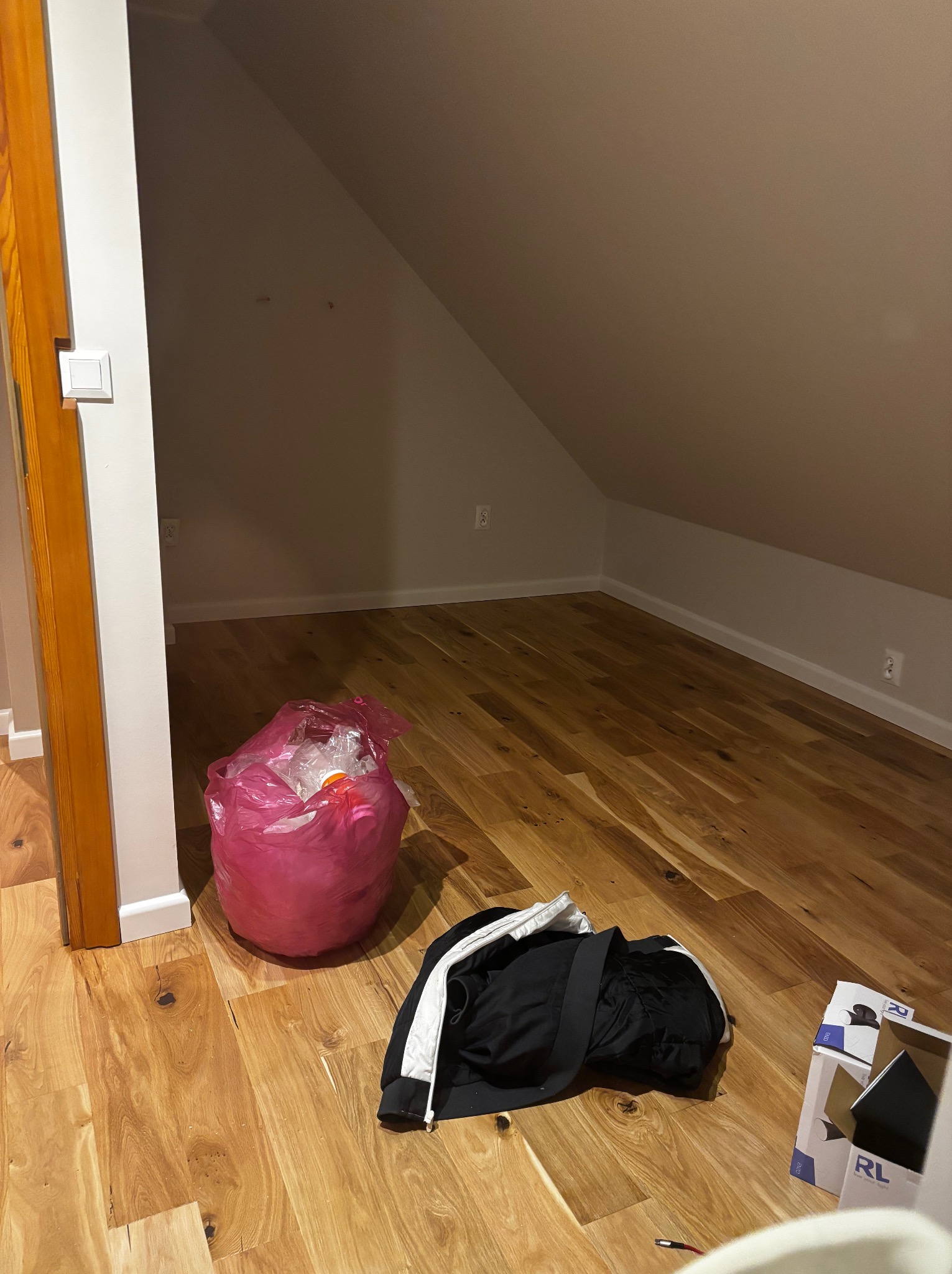 Jasna podłoga z paneli drewnianych w pokoju na poddaszu, widoczny fragment ściany, torba na śmieci i kartony. Ujęcie z niskiego kąta.