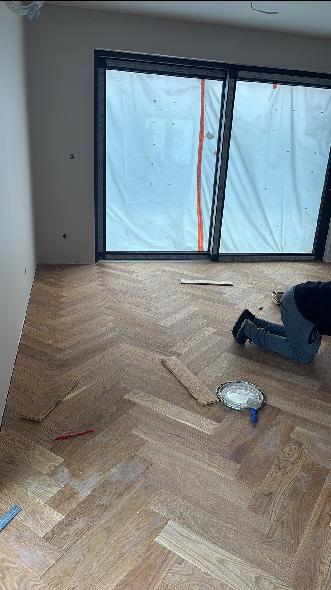 Układanie parkietu w jodełkę: widok z góry na częściowo ułożoną podłogę, narzędzia pracy i osobę w trakcie montażu. Okno zasłonięte folią ochronną.