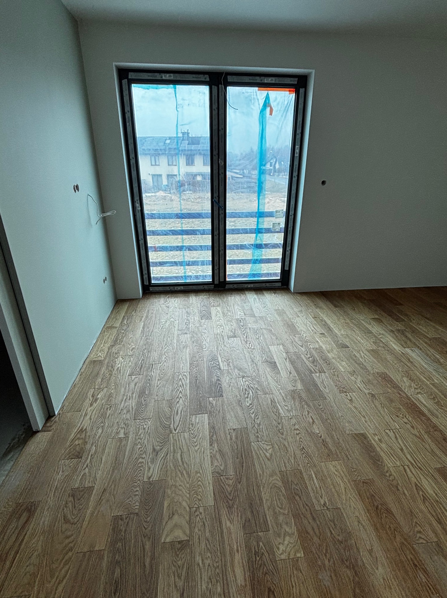 Ułożone panele podłogowe w jasnym pokoju z dużym oknem balkonowym zabezpieczonym folią. Widoczne wystające kable elektryczne. Podłoga w odcieniach brązu z wyraźnym wzorem drewna.
