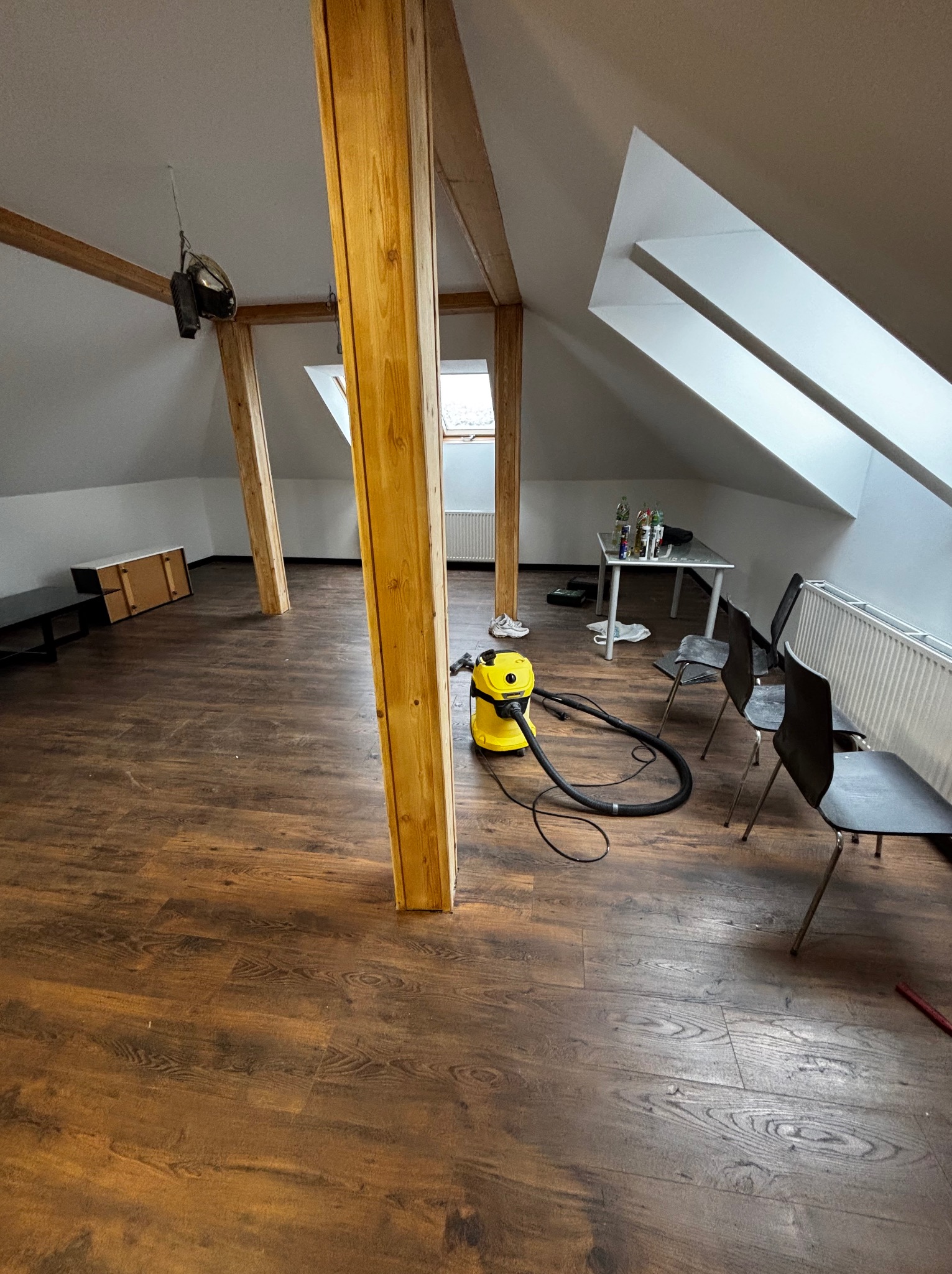 Poddasze z drewnianymi belkami i nową, ciemną podłogą. Żółty odkurzacz stoi na panelach. Widoczne okna dachowe i białe ściany. Ustawione krzesła i stół.