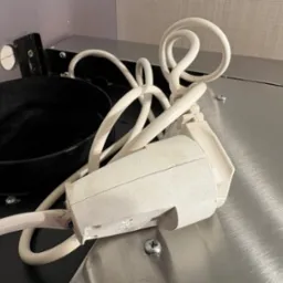 Brudna końcówka kabla zasilającego obok czarnej, okrągłej podstawki na metalowej powierzchni. Widoczne ślady użytkowania i zabrudzenia.