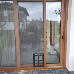 EKSPERT ROLET SPÓŁKA Z OGRANICZONĄ ODPOWIEDZIALNOŚCIĄ - Przesuwne drzwi balkonowe z moskitierą w drewnianej ramie. Widoczny fragment tarasu z szarych płyt i czarna klapka dla zwierząt. W tle dom z ogrodem.