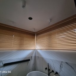 EKSPERT ROLET SPÓŁKA Z OGRANICZONĄ ODPOWIEDZIALNOŚCIĄ - Drewniane żaluzje poziome w łazience z marmurowymi ścianami i wolnostojącą wanną. Ujęcie z dołu, widoczne oświetlenie sufitowe.