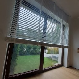 STOLICA ROLET SPÓŁKA Z OGRANICZONĄ ODPOWIEDZIALNOŚCIĄ - Białe żaluzje poziome zamontowane w oknie z widokiem na zielony ogród. Drewniana rama okienna i jasna podłoga. Delikatne światło dzienne.