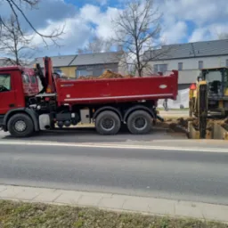Czerwona wywrotka z piaskiem obok wykopu zabezpieczonego szalunkiem, na tle zabudowy miejskiej i błękitnego nieba z chmurami. Prace ziemne w toku.