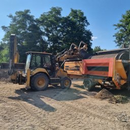 Trans-Janda s. c. Jakub Janda Bartosz Janda - Żółta koparko-ładowarka Doppstadt przesiewa ziemię na placu budowy w słoneczny dzień. Widoczne hałdy przesianej ziemi i drzewa w tle.