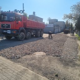 Trans-Janda s. c. Jakub Janda Bartosz Janda - Układanie żwiru na drodze: czerwona ciężarówka MAN z przodu, pracownik wyrównuje żwir, w tle koparka i ubijarka. Dzień słoneczny, niebieskie niebo.