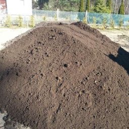 Trans-Janda s. c. Jakub Janda Bartosz Janda - Kupa ciemnej ziemi ogrodowej na trawniku, z widocznymi grudkami i nierównościami. W tle siatka ogrodzeniowa i drzewa.