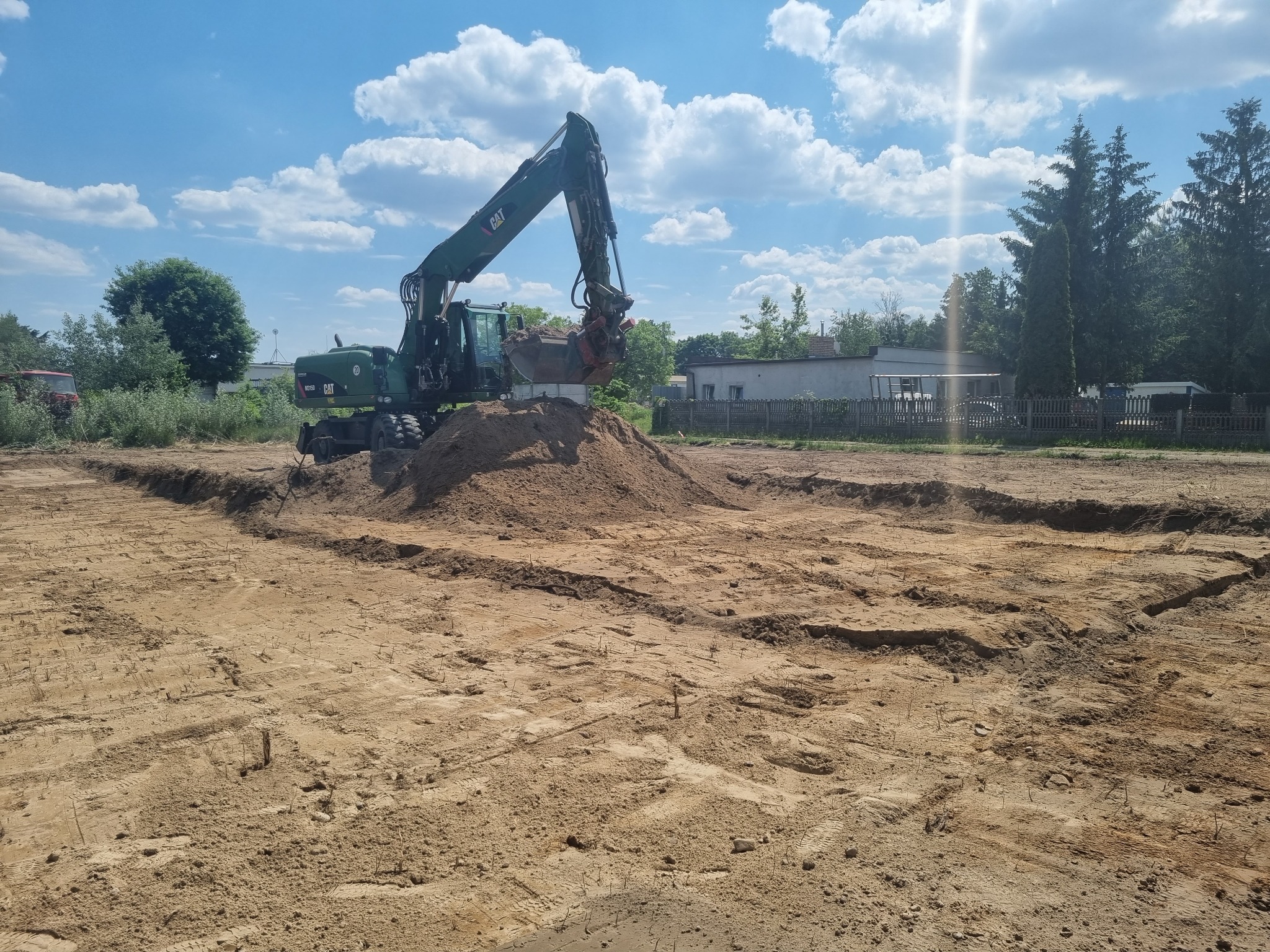 Zielona koparka CAT na placu budowy, usypująca górę piasku, widoczne wykopy. Błękitne niebo z chmurami w tle. Krajobraz po niwelacji terenu.
