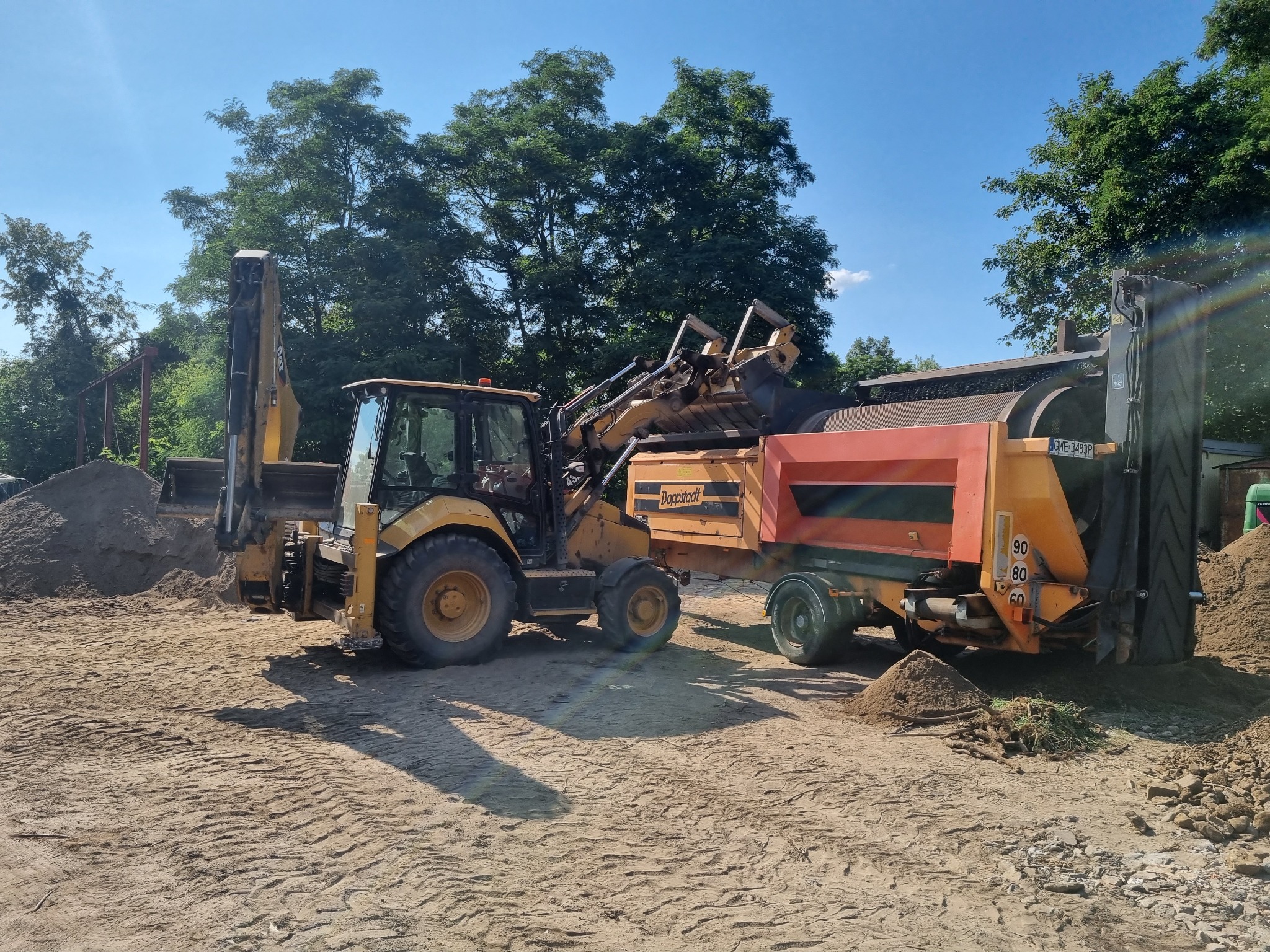 Żółta koparko-ładowarka Doppstadt przesiewa ziemię na placu budowy w słoneczny dzień. Widoczne hałdy przesianej ziemi i drzewa w tle.