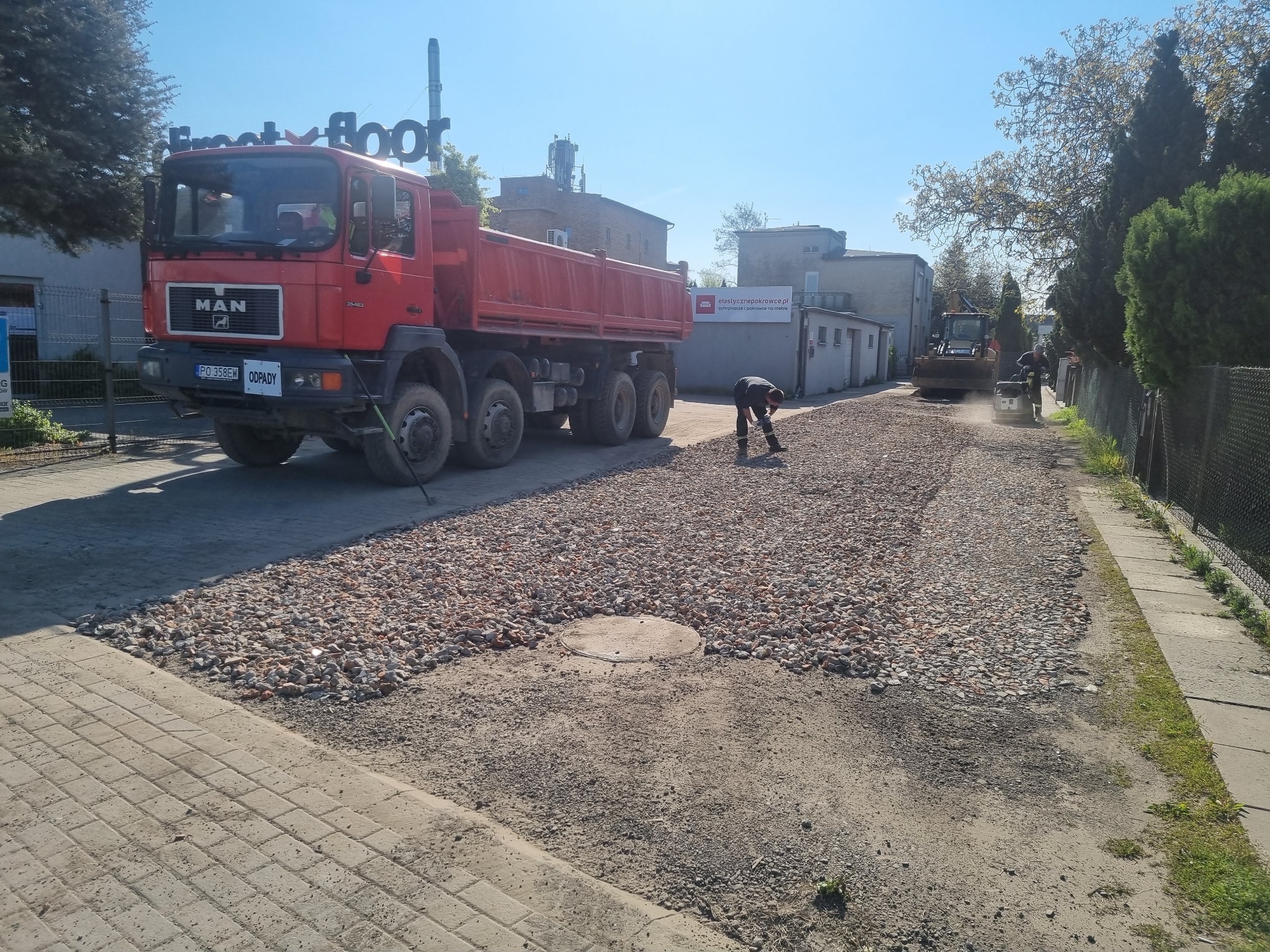Układanie żwiru na drodze: czerwona ciężarówka MAN z przodu, pracownik wyrównuje żwir, w tle koparka i ubijarka. Dzień słoneczny, niebieskie niebo.