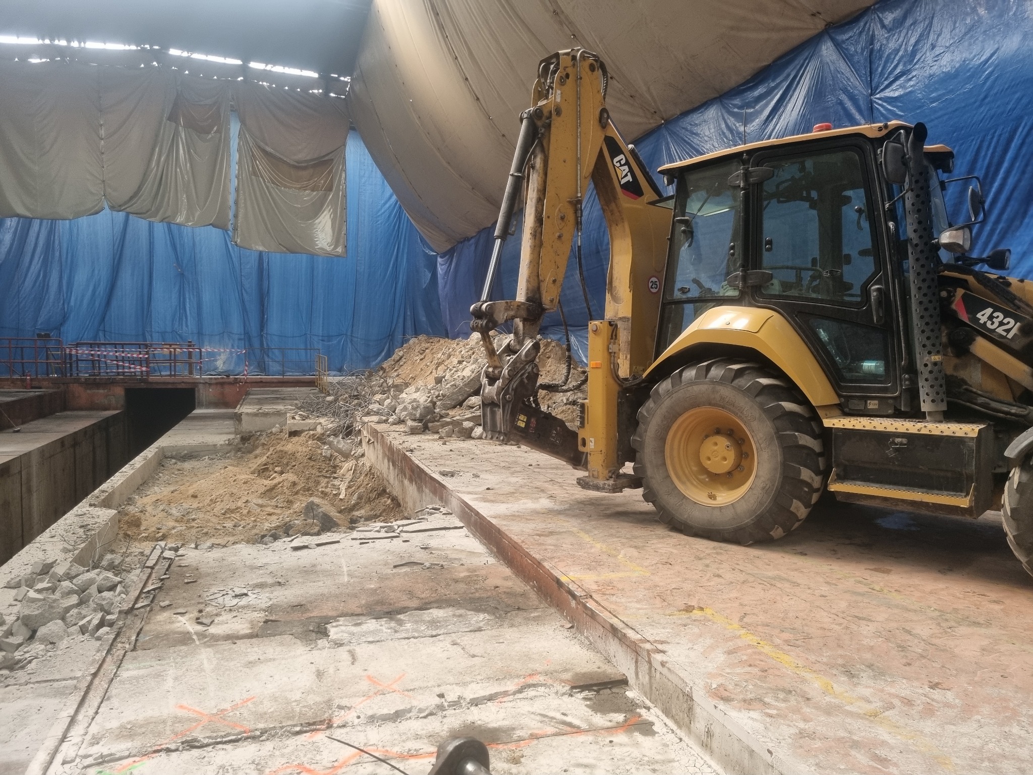 Żółta koparko-ładowarka CAT 432F2 na placu budowy w hali z niebieskim i białym płótnem w tle. Widoczny gruz i piasek. Etap prac ziemnych.