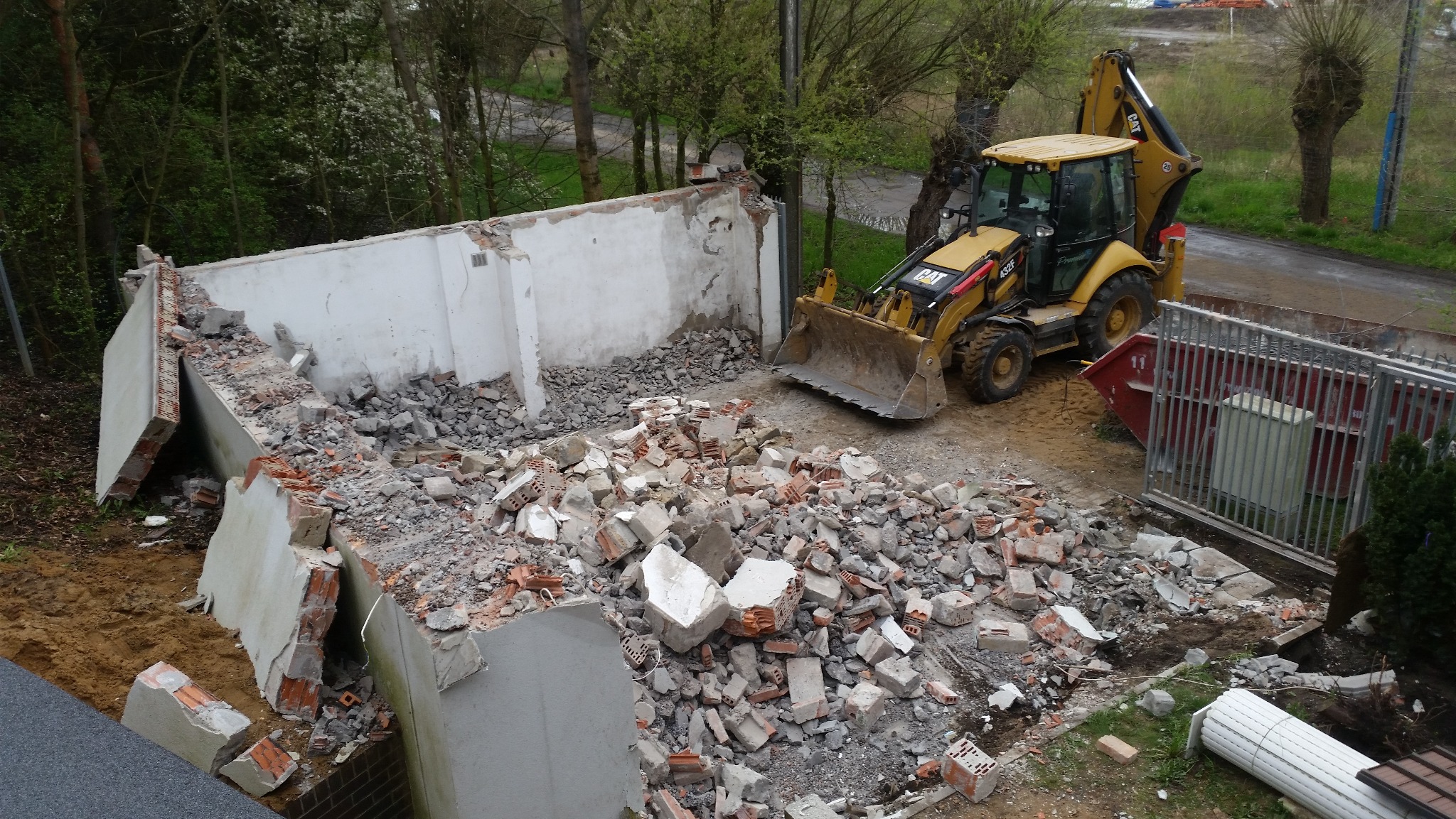 Częściowo zburzony budynek z gruzem i koparko-ładowarką CAT, widok z góry na plac rozbiórki z kontenerem na odpady w tle, zielone drzewa w otoczeniu.