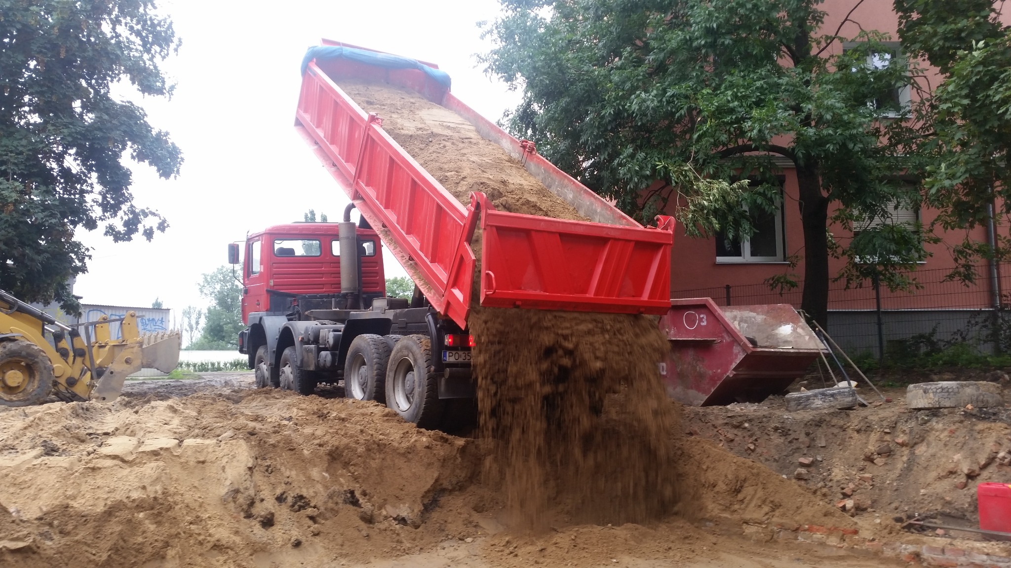 Czerwona wywrotka z uniesioną skrzynią wysypuje ziemię na plac budowy; w tle budynek i drzewa, z boku widoczna koparka i kontener. Ziemia w trakcie wysypywania.