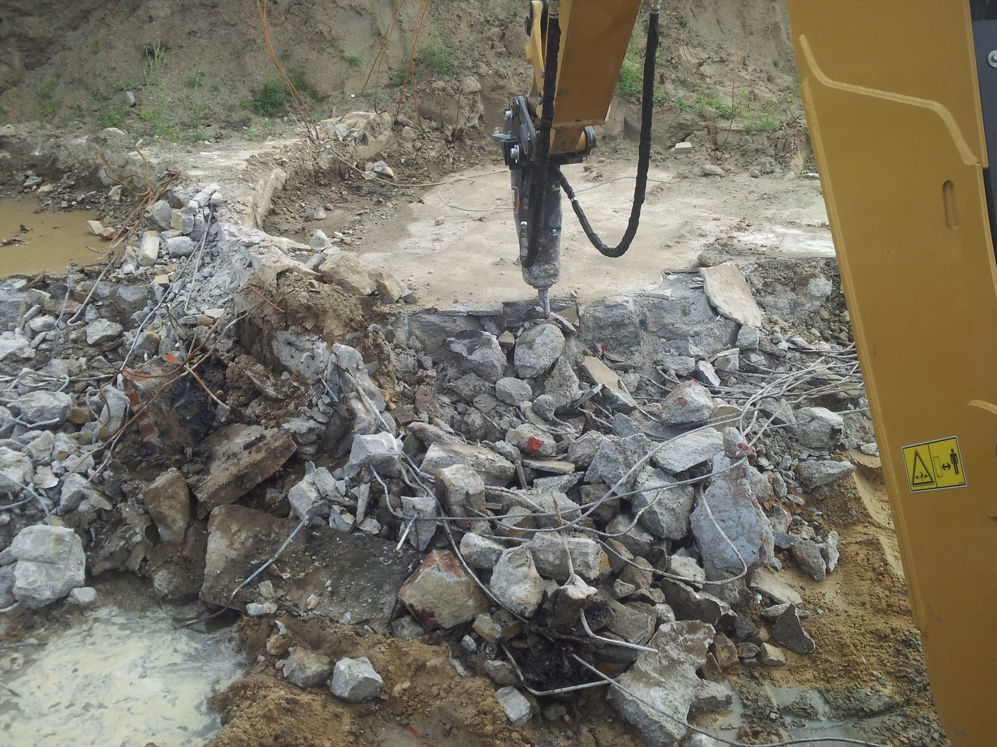Żółty młot hydrauliczny kruszy betonowe elementy na placu budowy. Fragmenty gruzu i ziemi w tle. Widoczne zbrojenia i resztki konstrukcji.