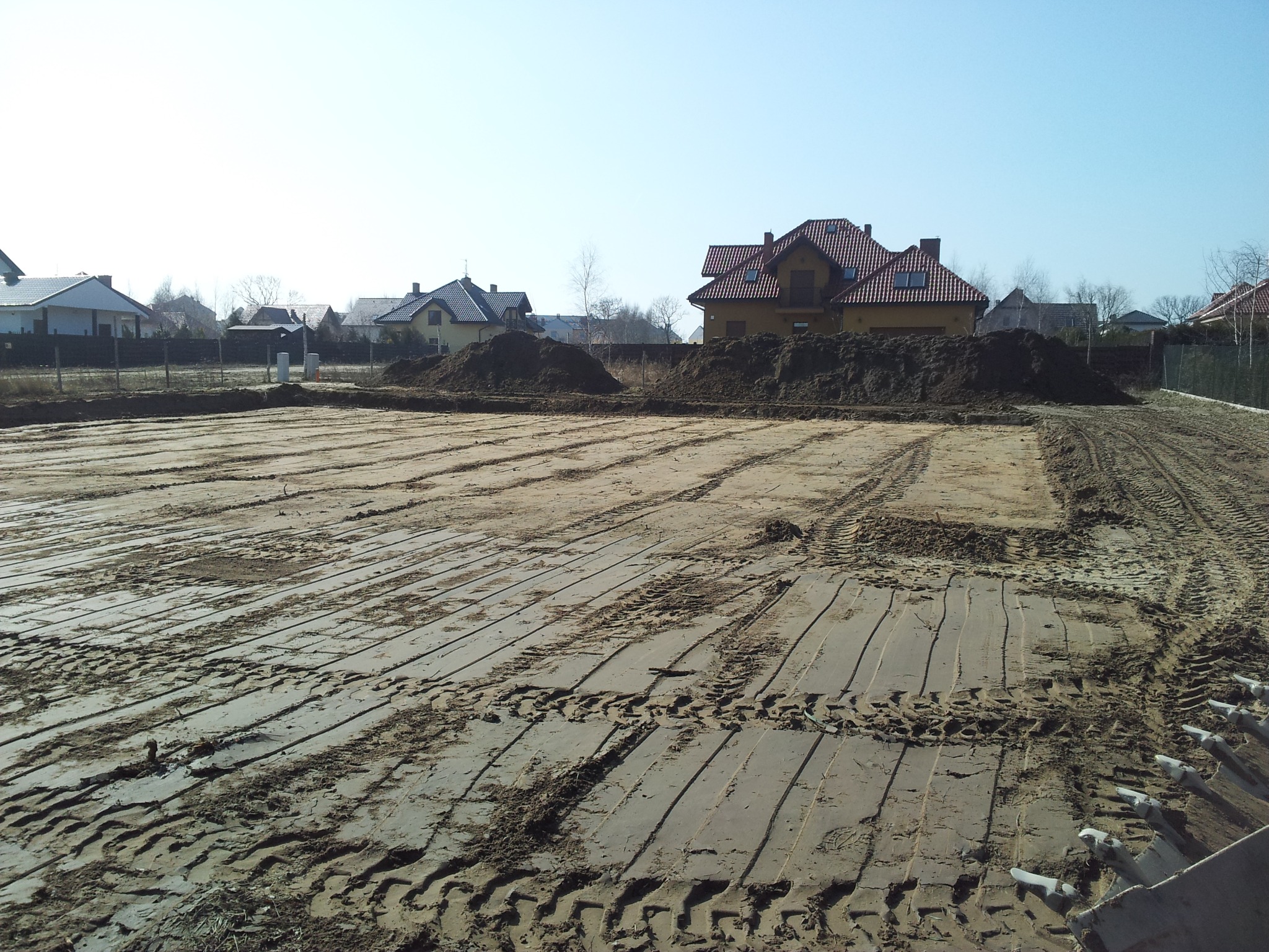 Przygotowany teren pod budowę z widocznymi śladami opon, w tle domy jednorodzinne i hałda ziemi. W oddali widoczne ogrodzenie z siatki.
