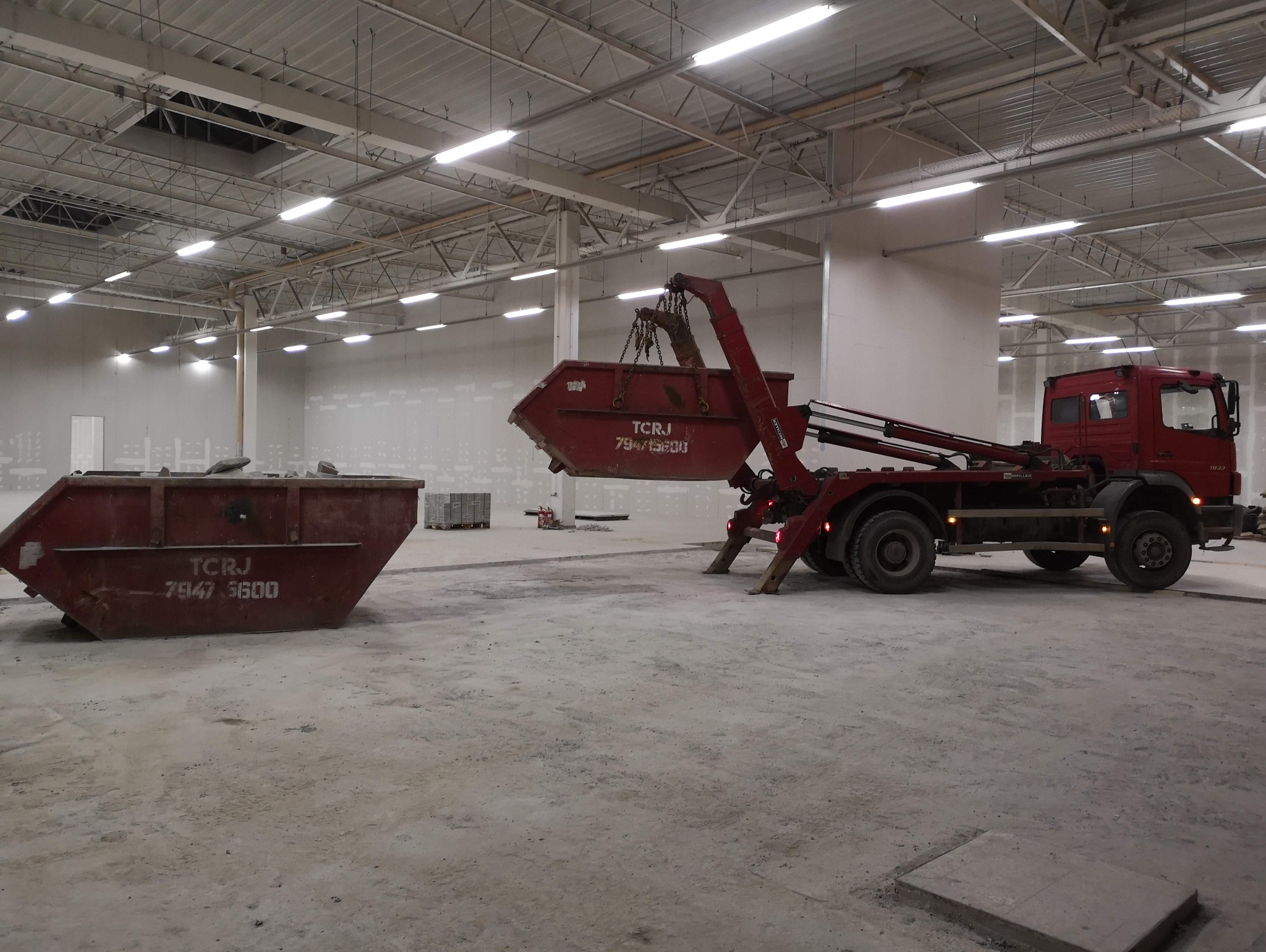 Ciężarówka z kontenerem na gruz w hali w Poznaniu. Kontener podnoszony, drugi stoi obok. Widoczne ślady po pracach budowlanych.