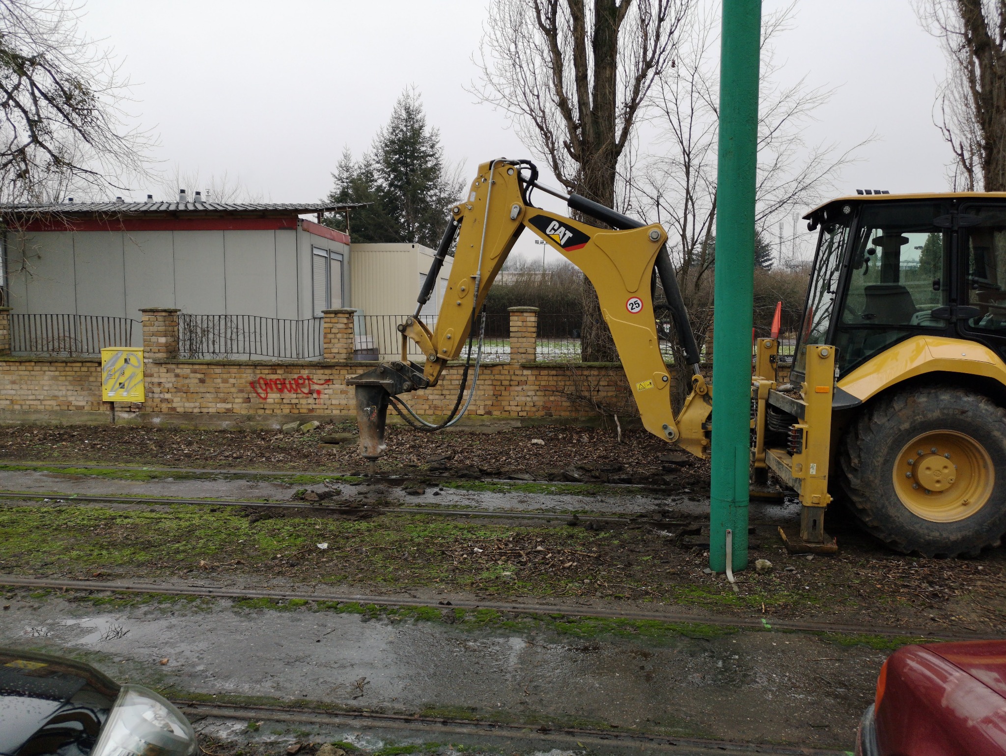 Żółta koparka CAT z młotem pneumatycznym rozbija tory tramwajowe na tle ceglanego muru i szarego budynku. Widoczny zielony słup i fragmenty aut.