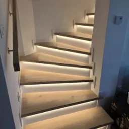 Drewniane schody z podświetleniem LED, widok z dołu, jasne drewno, białe ściany, nowoczesny design, oświetlenie punktowe na suficie, fragment poręczy.