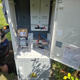 ADAM BREMER Activa Electric - Otwarta skrzynka elektryczna Jakmet na zewnątrz z widocznymi przewodami, bezpiecznikami i oznaczeniami. Częściowo widoczny pracownik w trakcie instalacji.