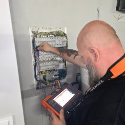 ADAM BREMER Activa Electric - Elektryk z tatuażem sprawdza napięcie w skrzynce bezpiecznikowej za pomocą miernika Sonel. Widoczne przewody i elementy instalacji elektrycznej.