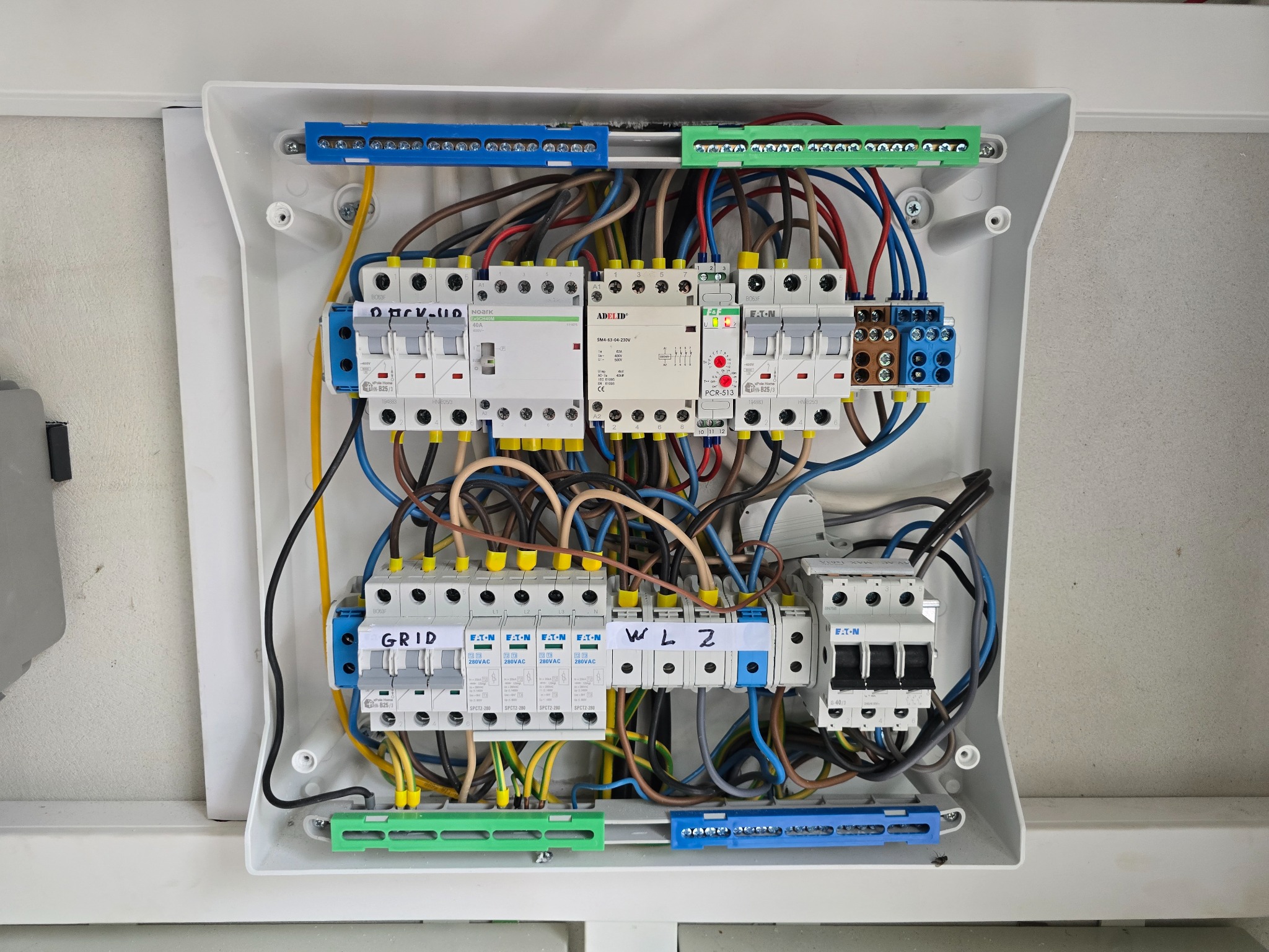 Rozdzielnica elektryczna w białej obudowie z widocznymi bezpiecznikami, przewodami i listwami zaciskowymi. Napisy 'GRID' i 'WL2' na elementach rozdzielnicy.