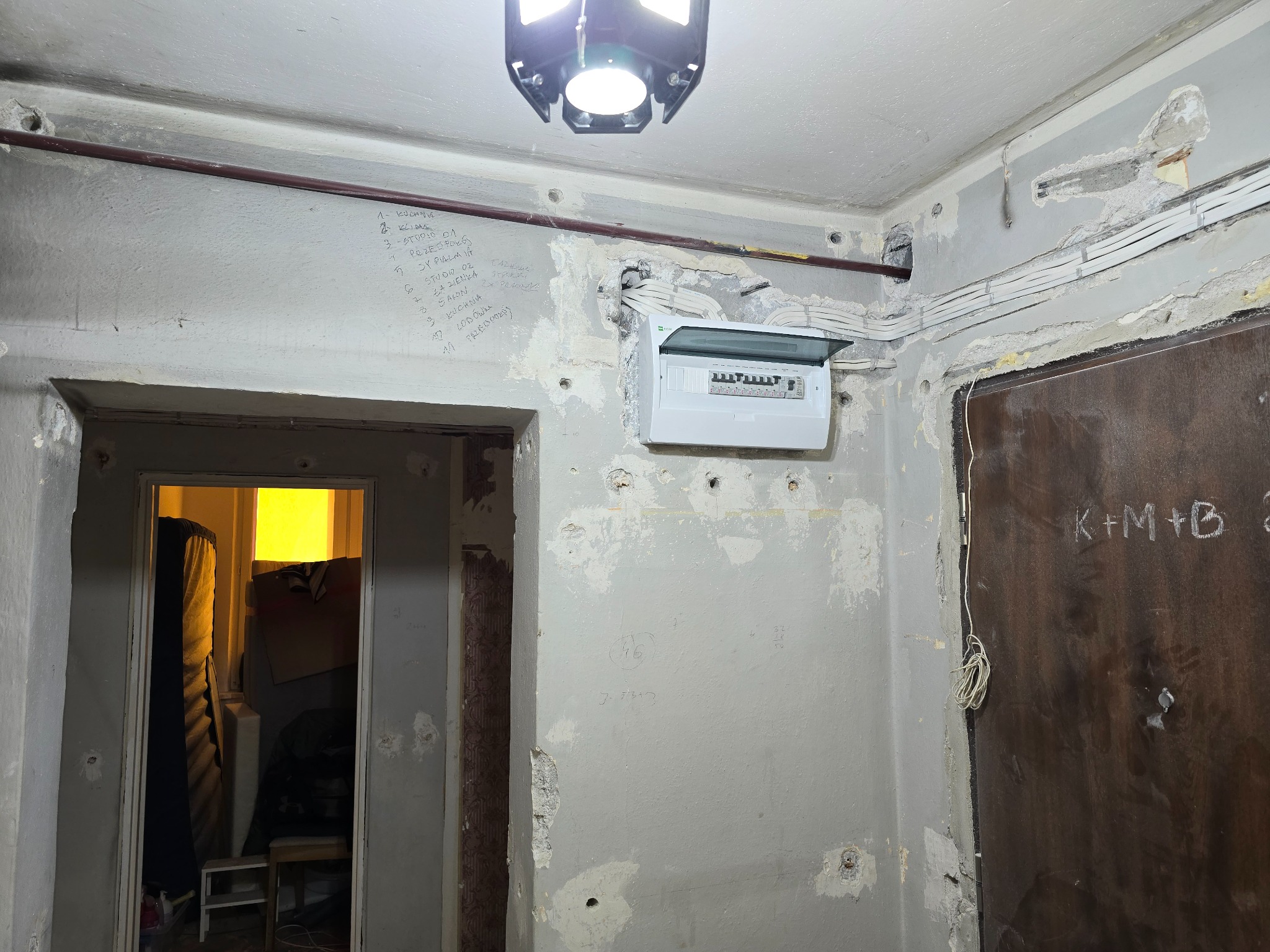 Rozdzielnica elektryczna z otwartą obudową na ścianie w trakcie remontu; widoczne przewody, ślady po kuciu i notatki na ścianie, surowy stan wykończenia.