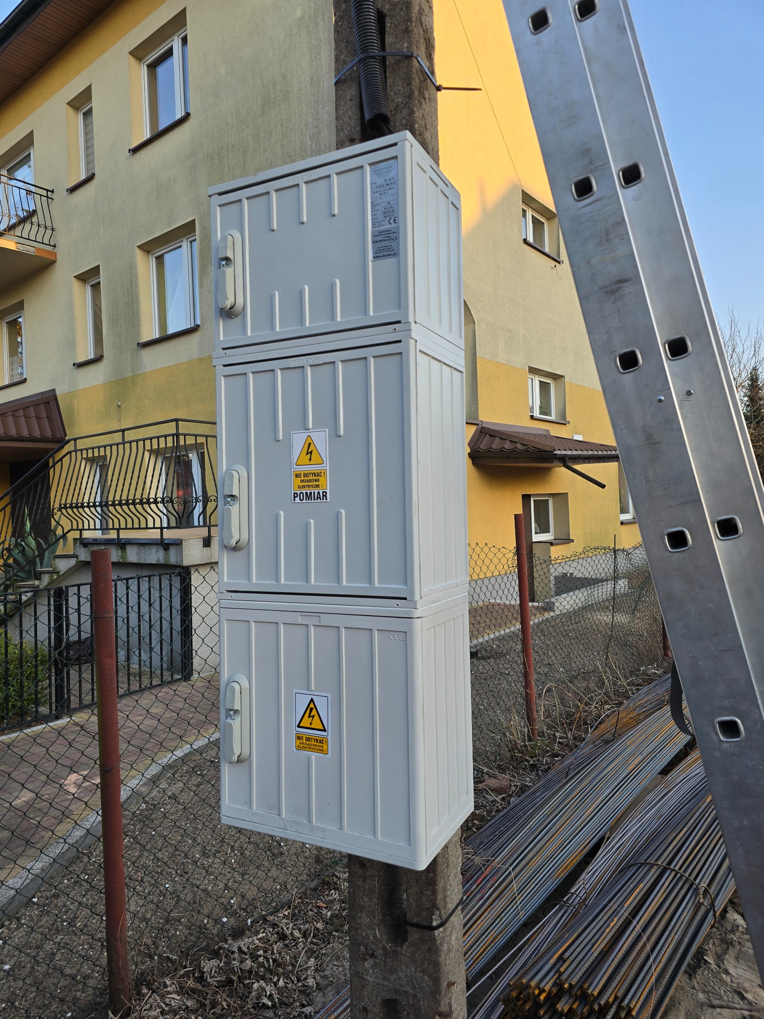 Szafka pomiarowa na słupie, z naklejkami ostrzegawczymi 'Nie dotykać! Urządzenie elektryczne! Pomiar'. Obok oparta drabina, w tle budynek mieszkalny.