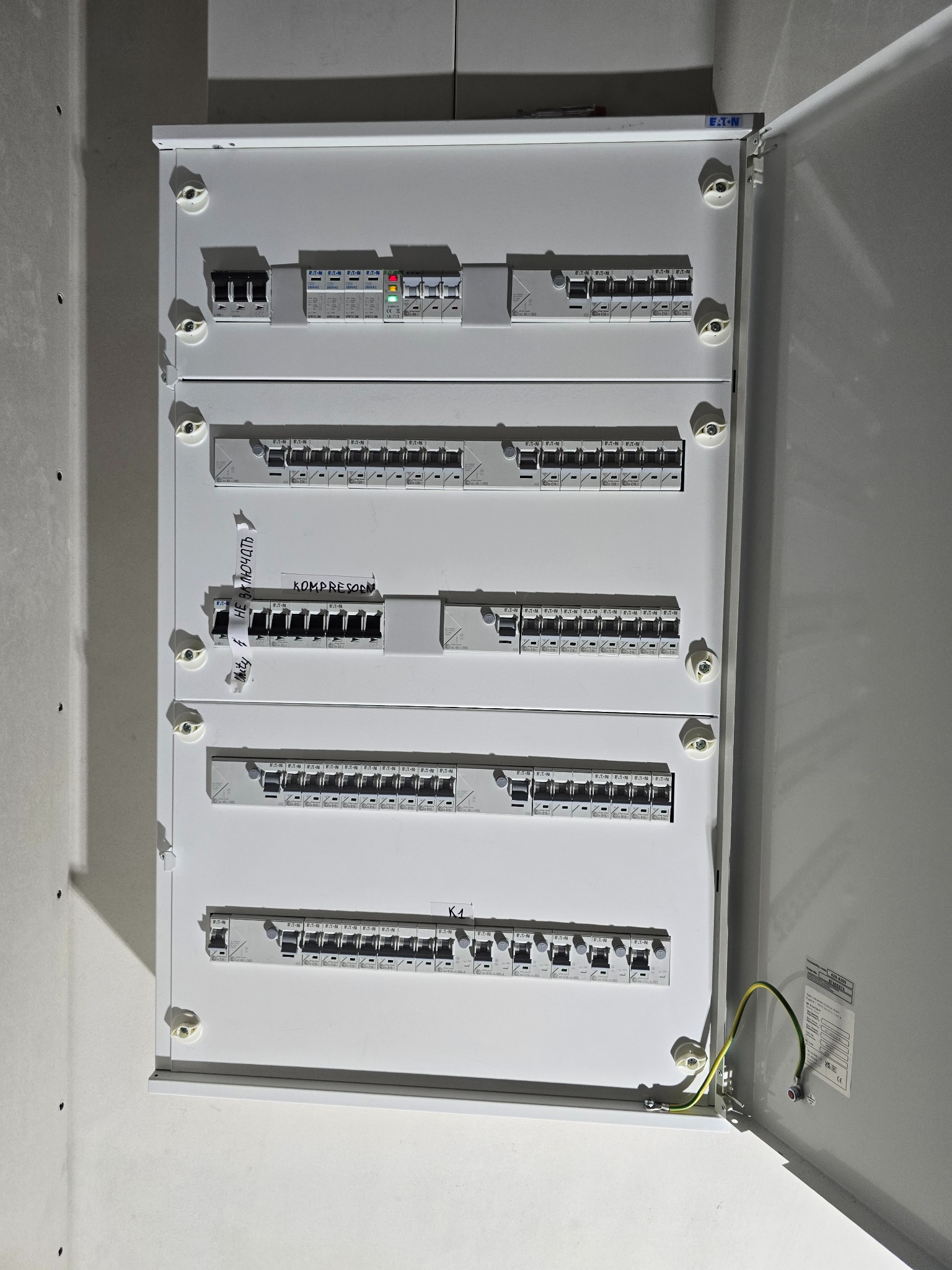 Otwarta, biała skrzynka elektryczna Eaton z bezpiecznikami i naklejkami z opisami obwodów. Widoczne przewody uziemiające i fragment ściany.