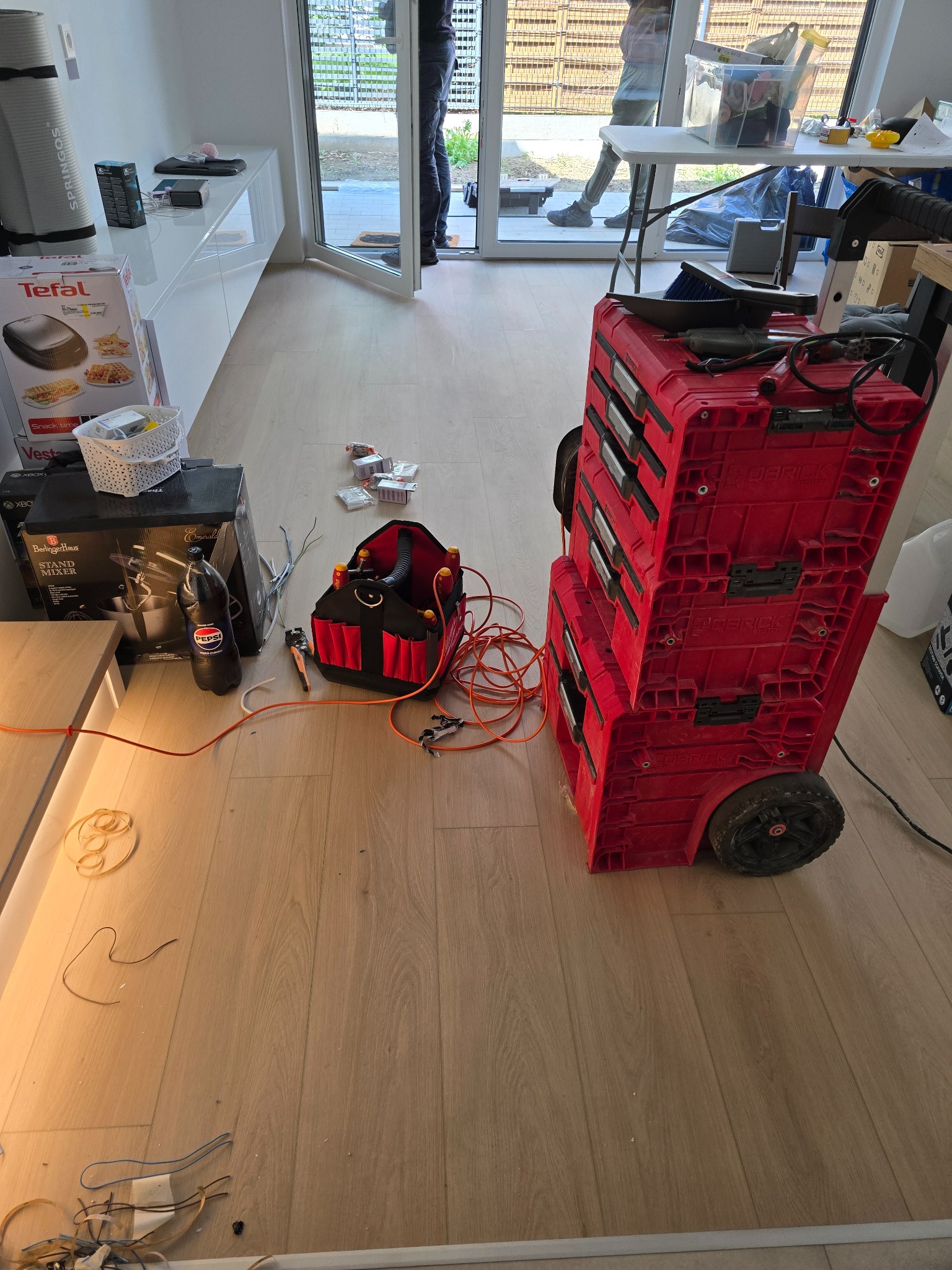 Rozłożone narzędzia elektryczne i skrzynka narzędziowa na podłodze w pomieszczeniu, kable i akcesoria widoczne w nieładzie. Skrzynka narzędziowa na kółkach.