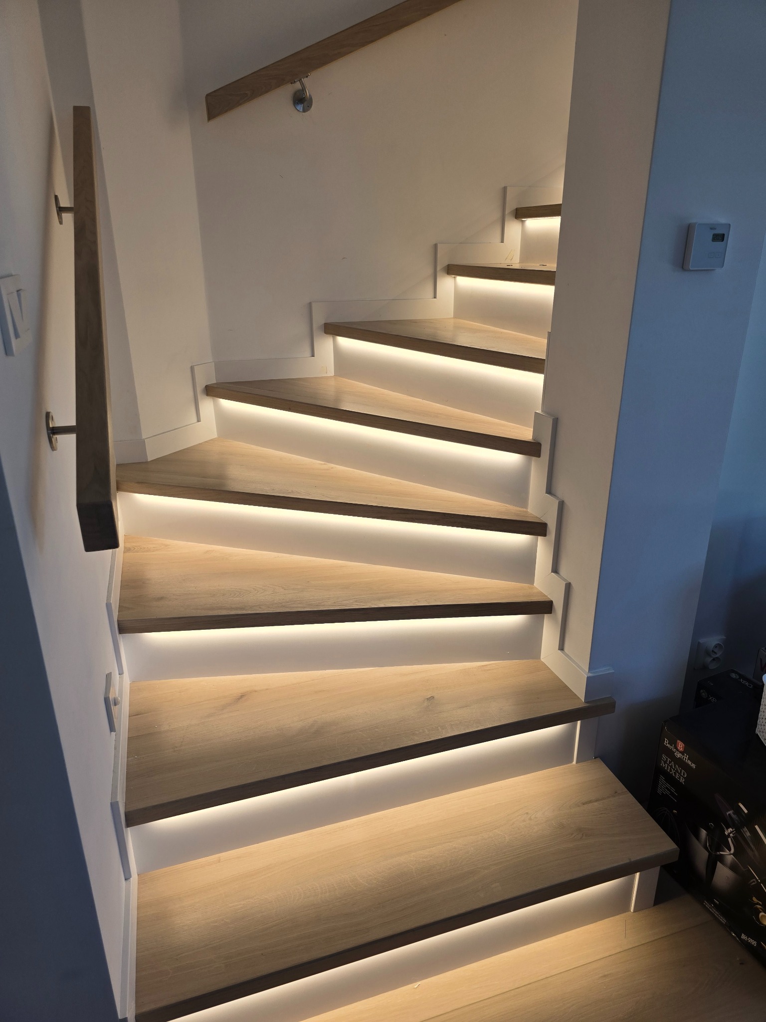 Drewniane schody z podświetleniem LED, widok z dołu, jasne drewno, białe ściany, nowoczesny design, oświetlenie punktowe na suficie, fragment poręczy.