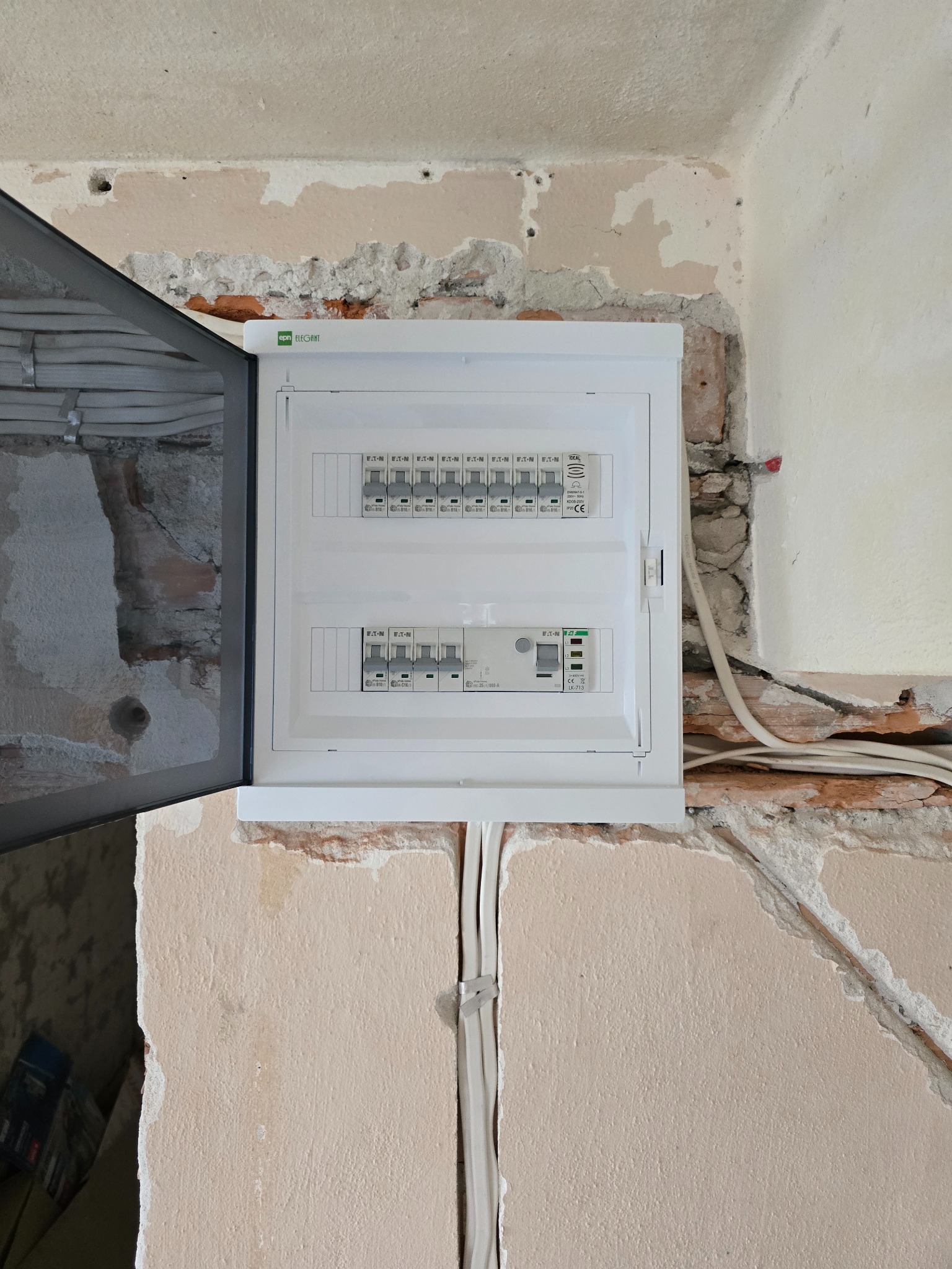 Rozdzielnia elektryczna EATON w trakcie montażu, widoczne kable i surowy stan ściany. Otwarta obudowa ukazuje zabezpieczenia.