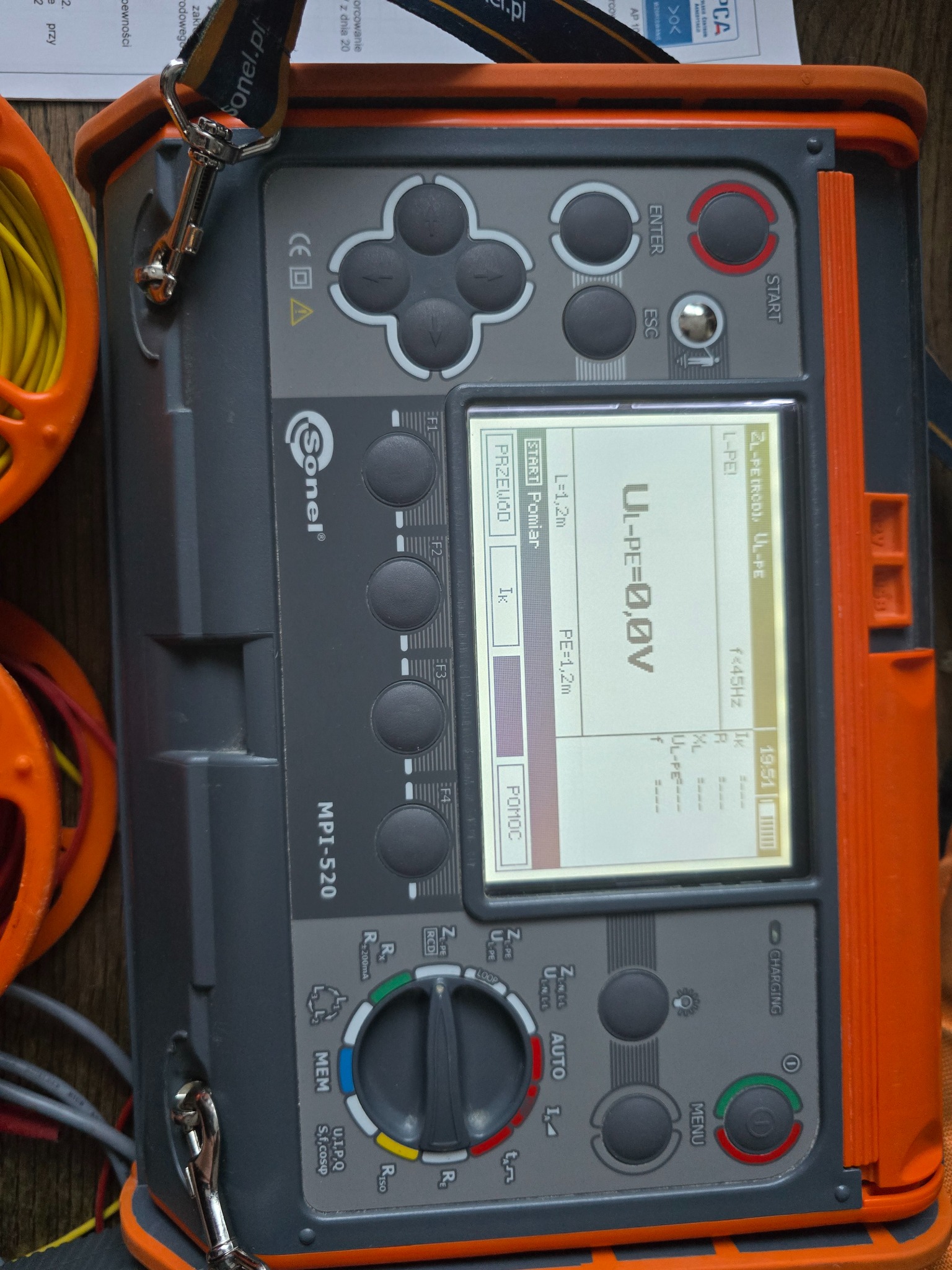 Miernik Sonel MPI-520 z wyświetlaczem wskazującym napięcie U-PE=0,0V, obok widoczne kable pomiarowe i fragment dokumentu. Urządzenie w obudowie szaro-pomarańczowej.