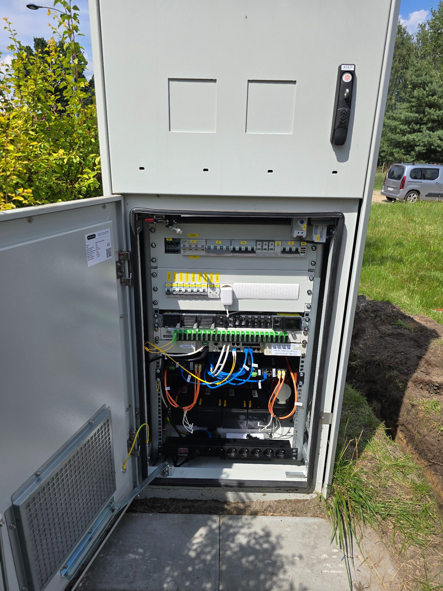 Otwarta szafa elektryczna z widocznymi zabezpieczeniami, okablowaniem i listwami. Na zewnątrz trawa i drzewa. Szafa na betonowej podstawie.