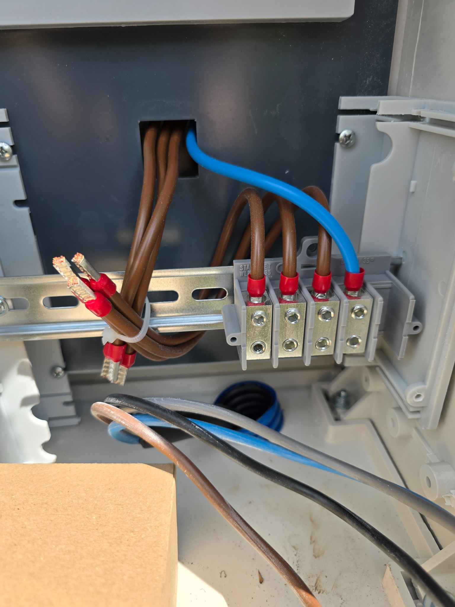 Wnętrze skrzynki elektrycznej z podłączonymi przewodami: brązowe, niebieskie, czarne i szare. Złącza kablowe z zaciskami śrubowymi. Widoczne detale mocowania przewodów.