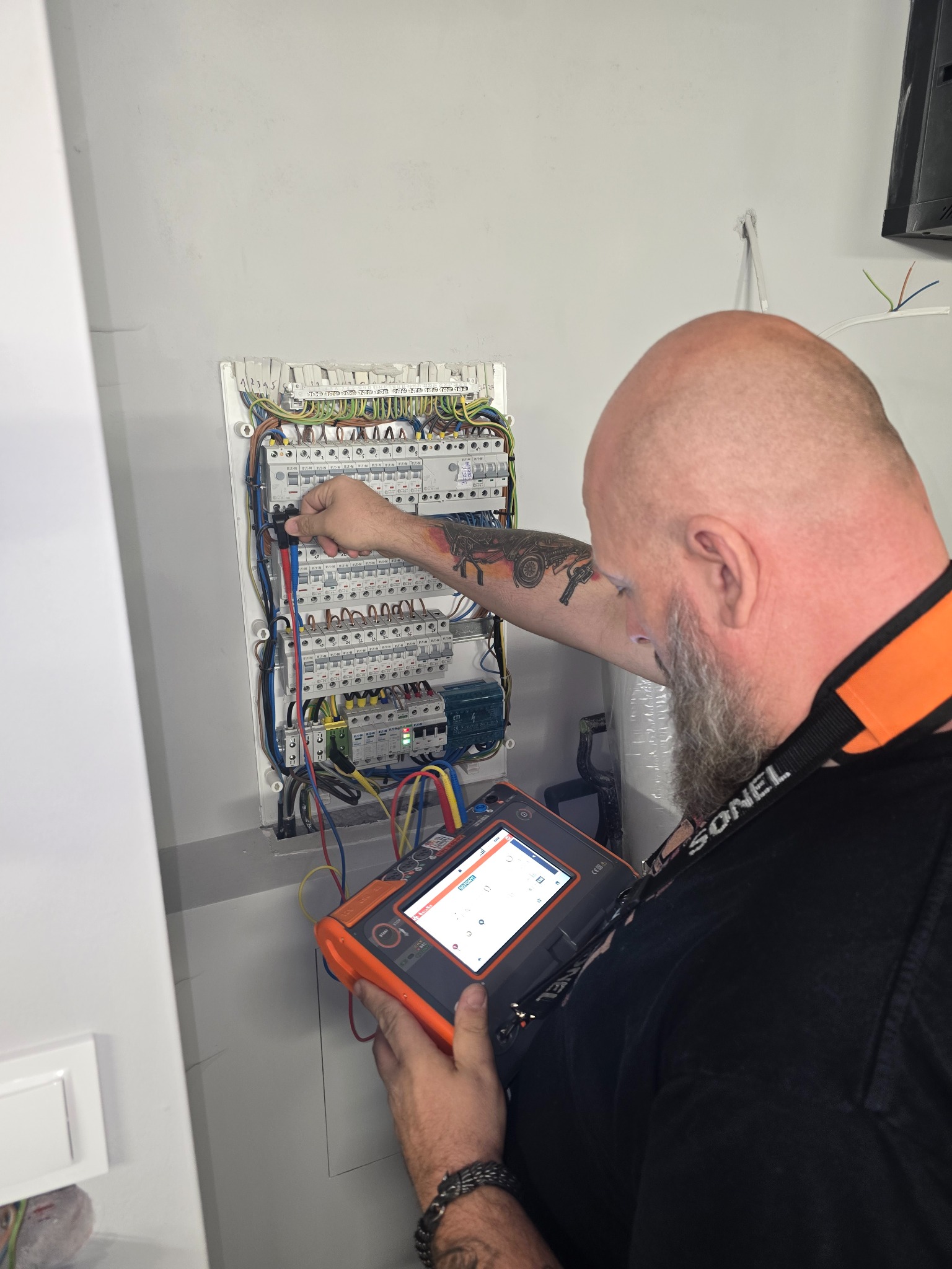 Elektryk z tatuażem sprawdza napięcie w skrzynce bezpiecznikowej za pomocą miernika Sonel. Widoczne przewody i elementy instalacji elektrycznej.