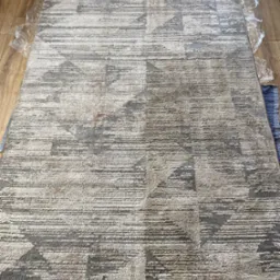 Wyczyszczony beżowo-szary dywan z geometrycznym wzorem, zabezpieczony folią na brzegach, leży na drewnianej podłodze. Widoczne ślady po czyszczeniu.